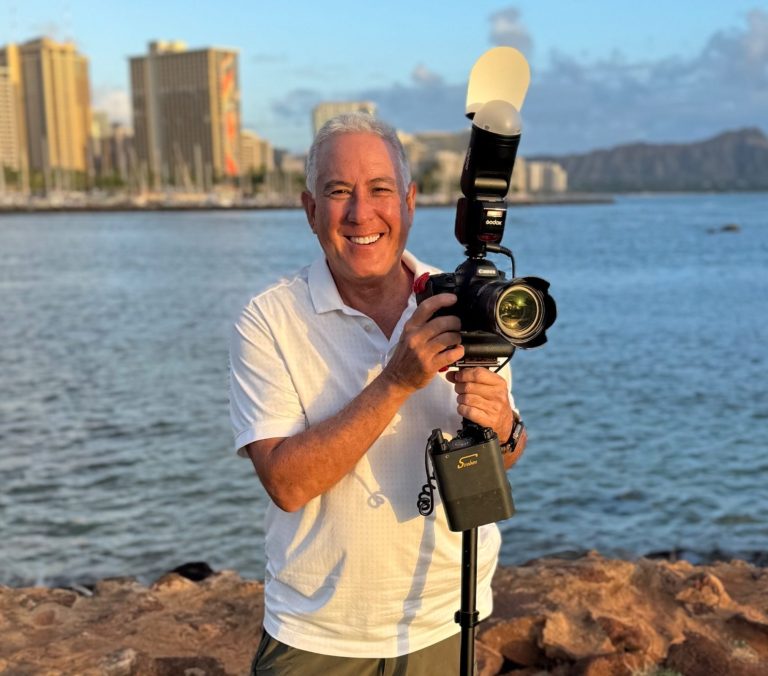 Hawaii beach weddings photographer holding a camera with a flash by the water and buildings in the background.