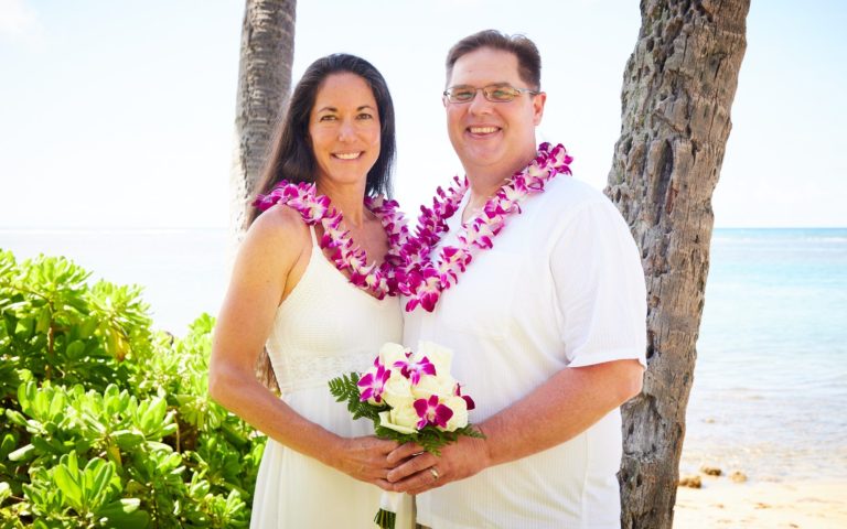 Hawaii Weddings at Kahala Beach. Smiling couple in white clothing wearing flower leis, holding a bouquet after their vow renewal at Kahala Beach.